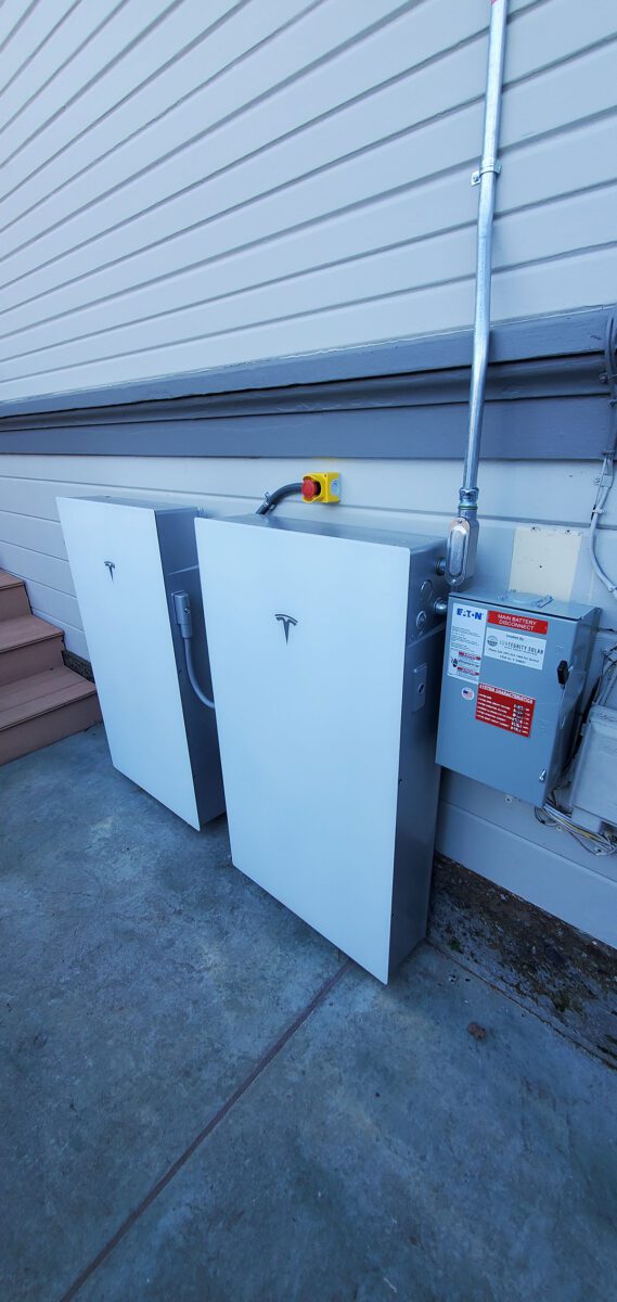 energy-storage11 Two white Tesla Powerwall battery units, part of a Santa Rosa solar battery energy storage setup, are mounted on an exterior wall beside a gray electrical panel and emergency shutoff switch, with conduit piping above on the concrete surface.