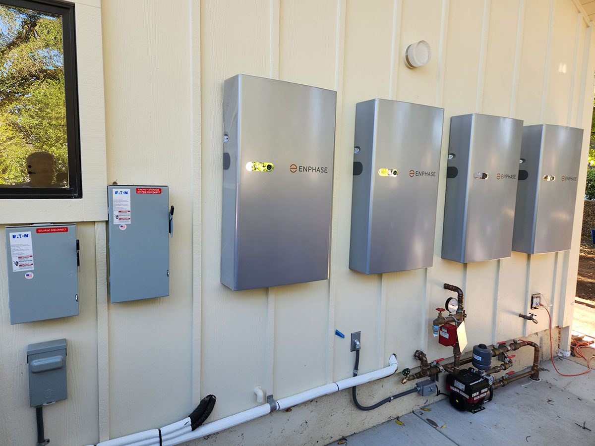 energy-storage12 Four large Enphase electrical panels and two smaller control boxes, part of a Santa Rosa solar battery energy storage setup, are mounted on an exterior wall, with pipes and utility equipment below and outdoor scenery reflected in a nearby window.