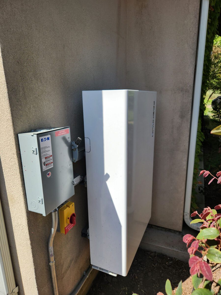 energy-storage13 A white rectangular battery storage unit, part of a Santa Rosa solar battery energy storage setup, is mounted on an exterior wall beside a gray electrical box. Some leaves from a nearby plant are visible in the lower right corner.