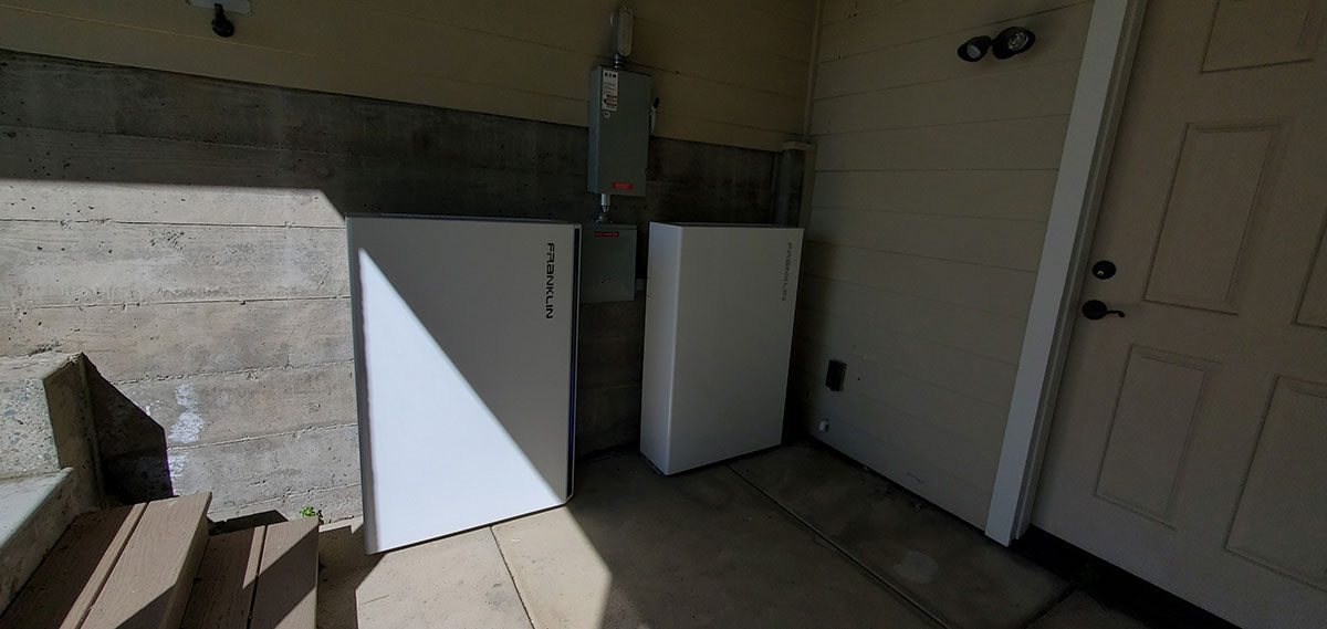 energy-storage2 Two white santa rosa solar battery energy storage units are installed against a wall next to an electrical panel, near outdoor steps and a closed door in a shaded area.