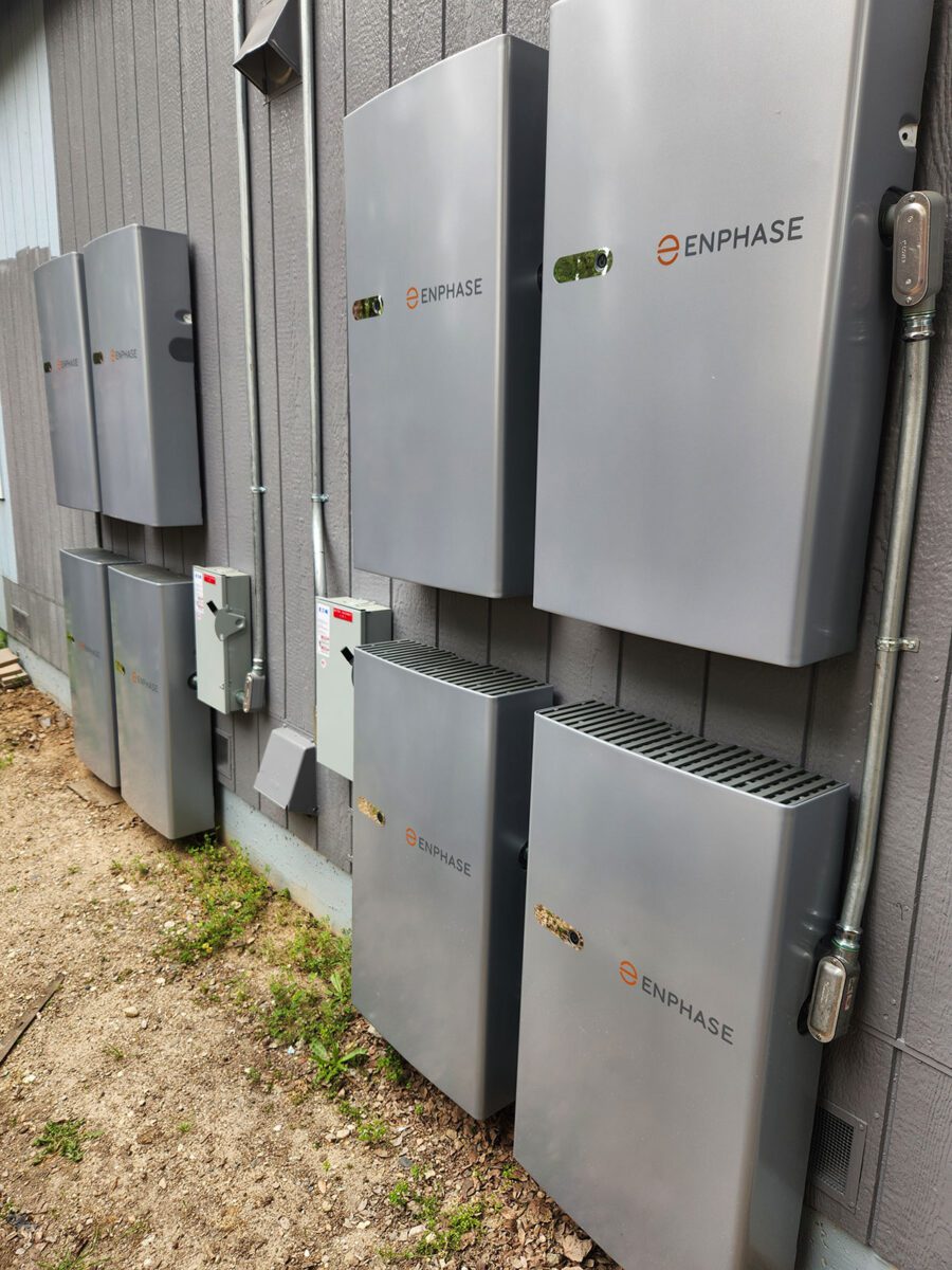 energy-storage3 Six large, gray Enphase battery storage units are mounted on the exterior wall of a building in Santa Rosa, showcasing solar battery energy storage; electrical panels and conduit are visible nearby, while the ground below is dirt with sparse grass.