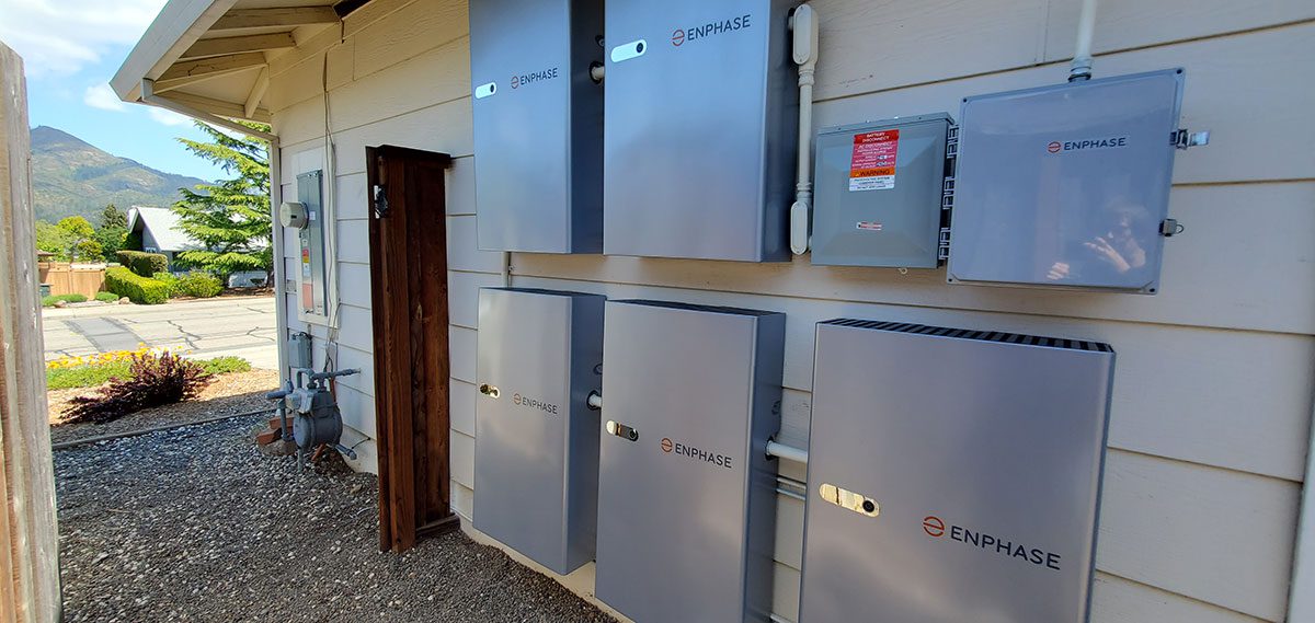 energy-storage4 Outdoor wall of a house in Santa Rosa with several large gray Enphase solar battery energy storage units and electrical boxes installed; trees and mountains are visible in the background.