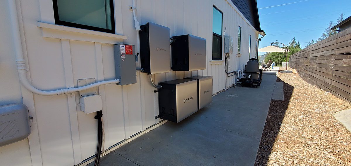 energy-storage6 Several santa rosa solar battery energy storage units are mounted on the exterior wall of a building beside a concrete walkway, with mulch and a wooden fence on the right. Outdoor equipment and trees are visible in the background.