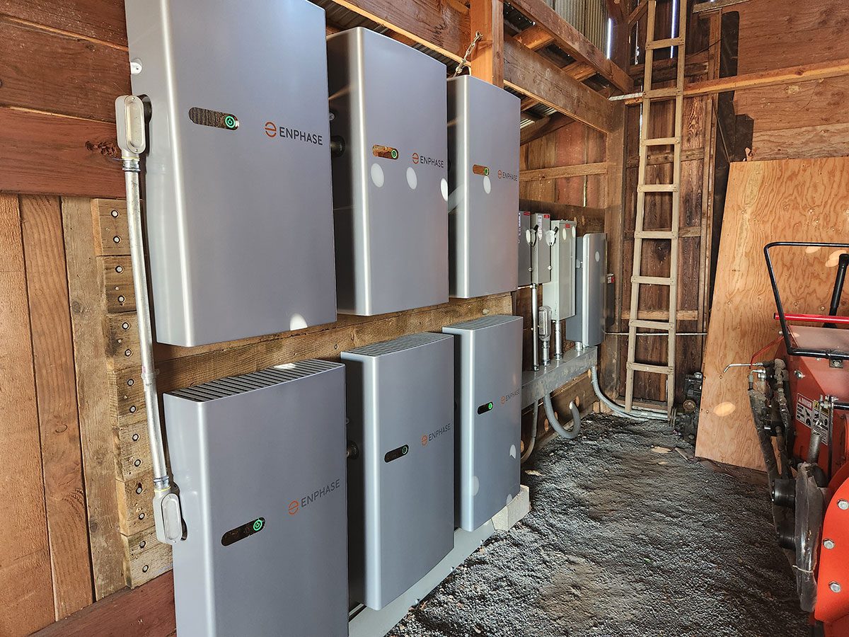 energy-storage7 Six metallic Enphase energy storage battery units, part of a Santa Rosa solar battery energy storage setup, are mounted on a wooden wall inside a shed, with visible wiring; a gravel floor and wooden ladder appear in the background.