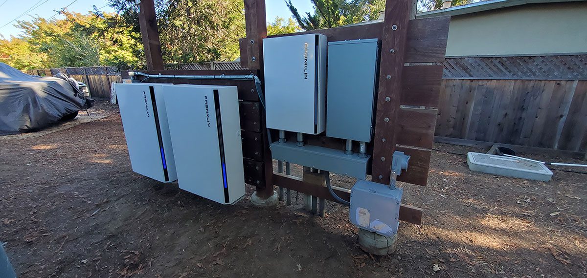 energy-storage8 Four Tesla Powerwall battery units and electrical boxes are mounted on wooden posts in a backyard near a fenced area and trees, showcasing a santa rosa solar battery energy storage setup. The ground is dirt, with a covered item to the left.