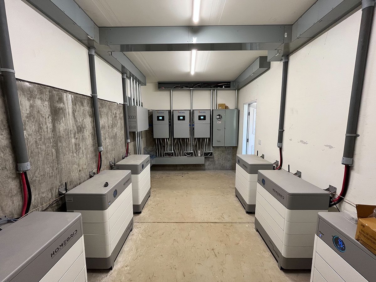 energy-storage9 A clean utility room in Santa Rosa with six large solar battery energy storage units along the walls, grey pipes running to the ceiling, and electrical panels on the far wall; the floor is bare concrete.
