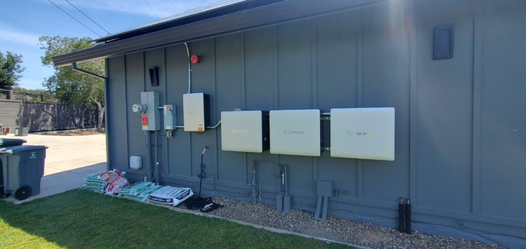 Three large white electrical units and a gray utility box are mounted on an exterior gray wall. Bags of garden soil or mulch are on the grass nearby, next to a driveway and trash bins.
