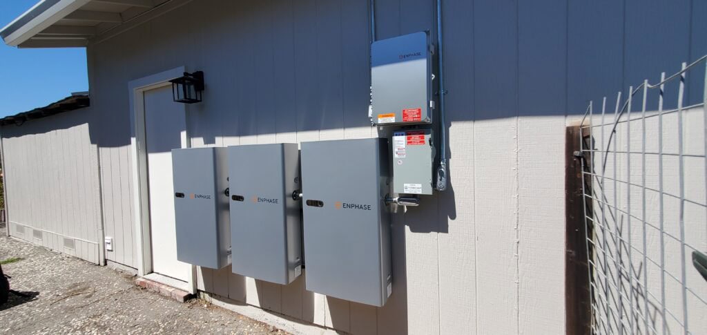 Three Enphase electrical panel boxes and an additional control box are mounted on the exterior wall of a light-colored building, next to a door and a metal fence, under a clear blue sky.