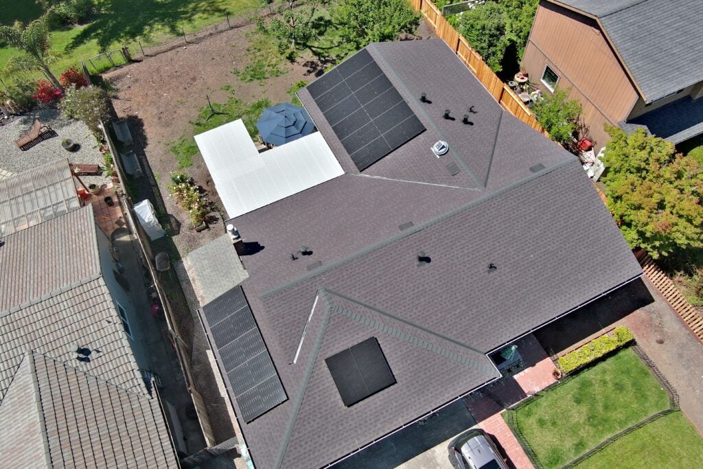 Aerial view of a house with dark roof shingles and solar panels installed. The backyard features a covered patio with a white roof and umbrella. Neighboring homes, fencing, and landscaping are also visible.