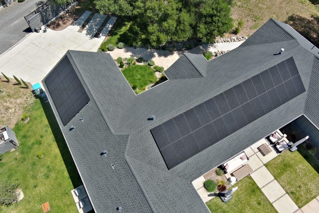 Aerial view of a house with a dark gray roof featuring solar panels, surrounded by green lawn, trees, a patio area with outdoor furniture, and a driveway with parked vehicles.
