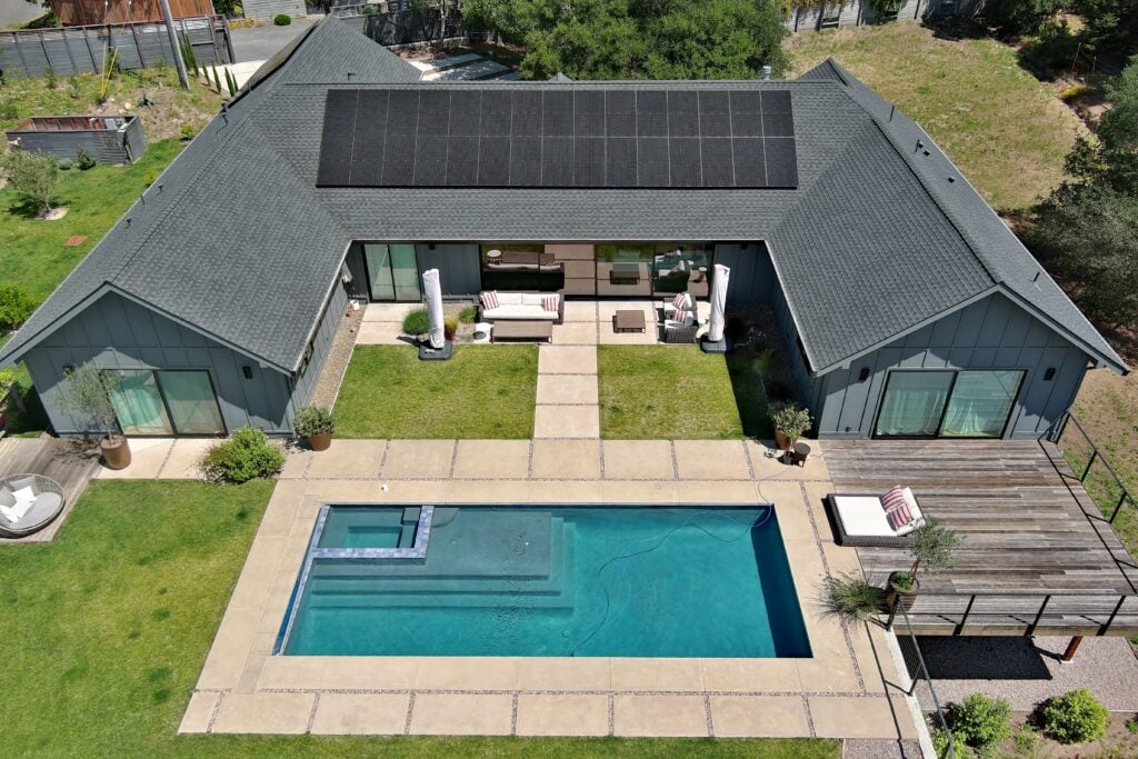 Aerial view of a modern house with dark roof solar panels, open patio, outdoor seating, and a rectangular pool with hot tub in a grassy backyard.