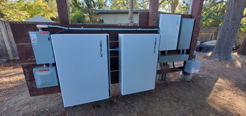 Two Franklin-branded electrical boxes and other equipment are mounted on a wooden structure outdoors, surrounded by trees, a fence, and a house in the background.