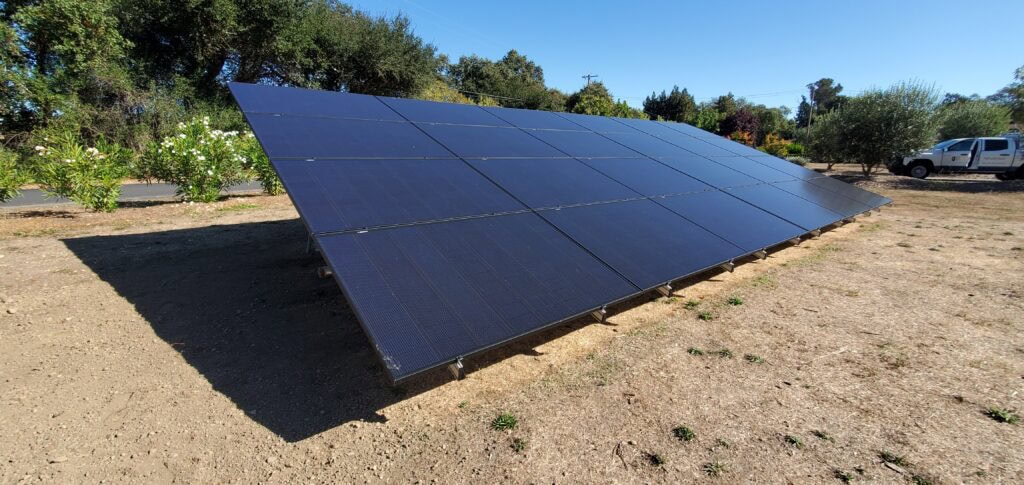 A large ground-mounted solar panel array is installed on dry, bare soil in a sunny outdoor area, with trees, bushes, and a parked truck in the background.