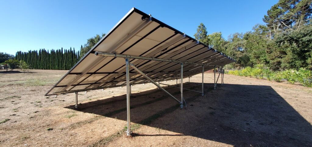 A ground-mounted solar panel array stands in a dry, open area with trees and a clear blue sky in the background.