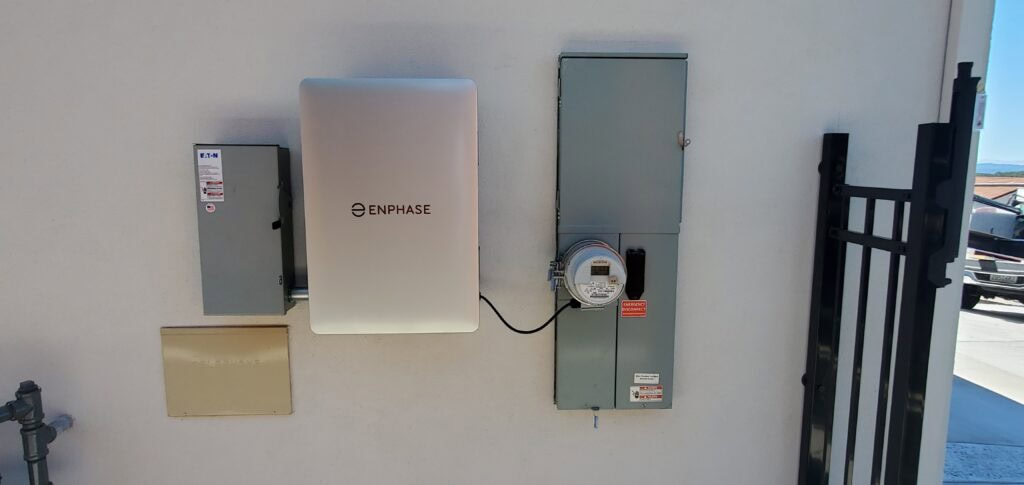 Enphase battery system, utility meter, and electrical panels mounted on an exterior white wall near a black metal gate in bright sunlight.