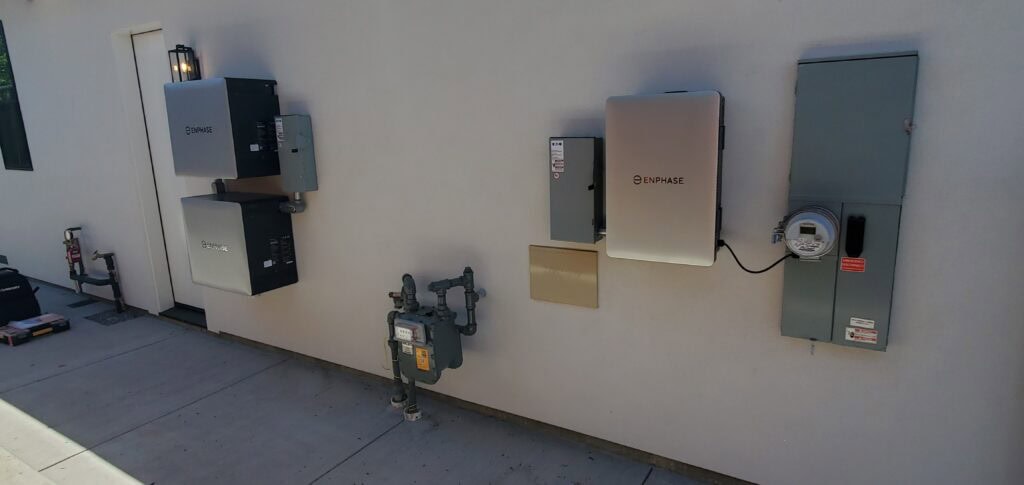 Two Enphase battery systems and multiple electrical panels are mounted on an exterior wall near a gas meter. The setup appears to be part of a home energy storage and distribution system.