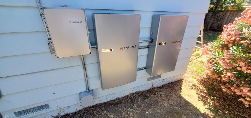 Three metal Enphase electrical panels are mounted on the exterior wall of a light blue house, near some bushes and a grassy patch.