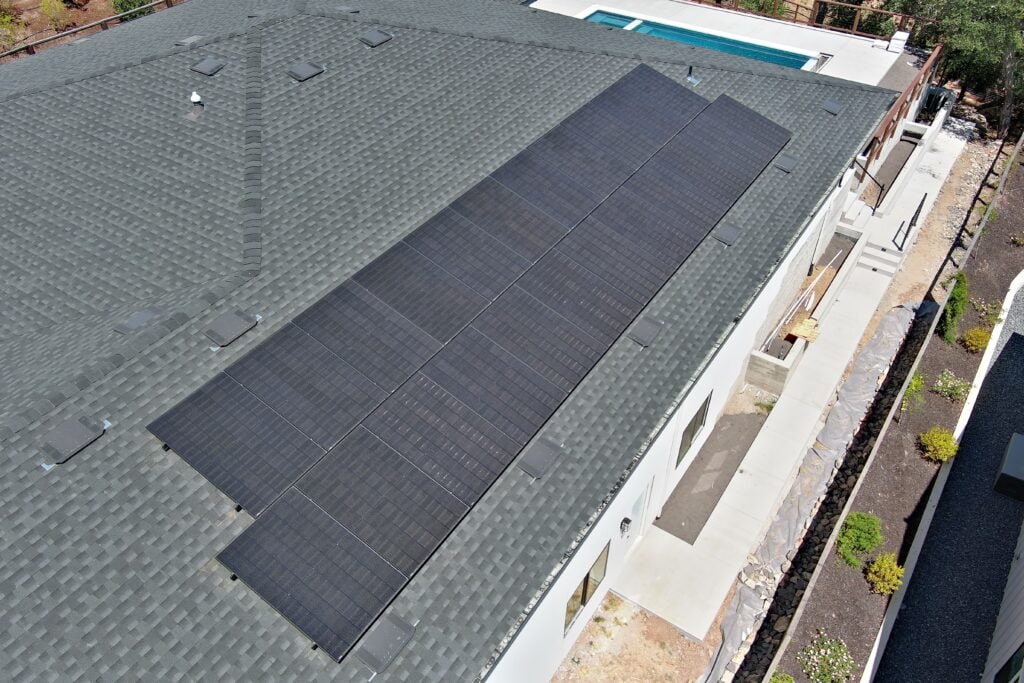 A row of black solar panels is installed on the gray shingled roof of a modern white house, with a pool and yard visible nearby.