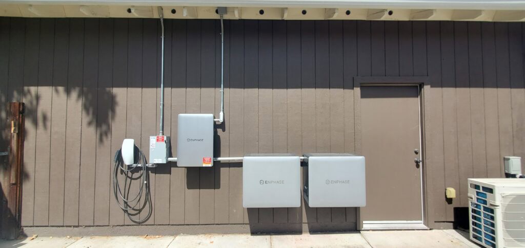 Two Enphase electrical boxes, conduit, a charger, and a utility meter are mounted on a brown wall next to a closed door, with an HVAC unit on the right and shadows from trees above.