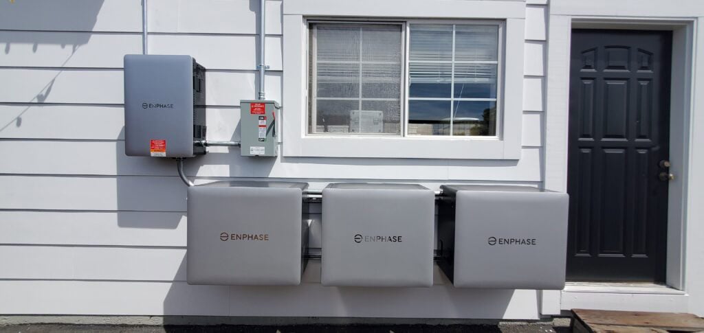 Three Enphase battery units and an electrical panel are mounted on the exterior wall of a house beneath a window, next to a black door. The wall is painted white with horizontal siding.