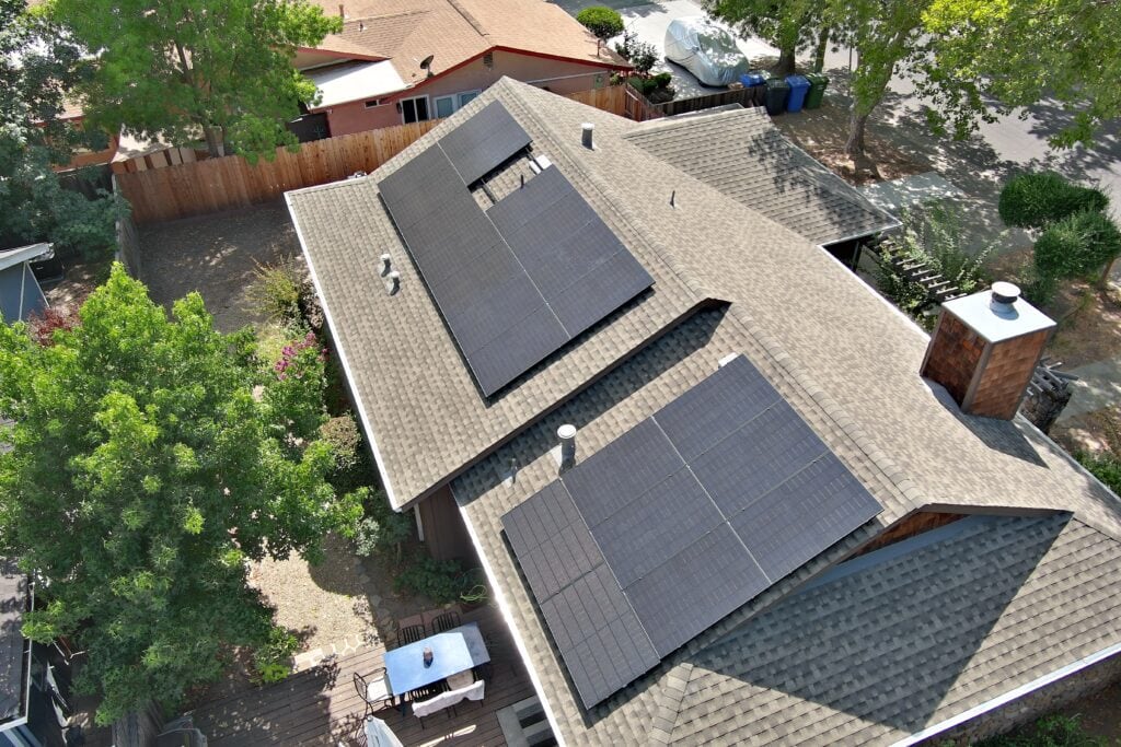 Aerial view of a house with solar panels installed on the roof, surrounded by trees and a fenced yard.