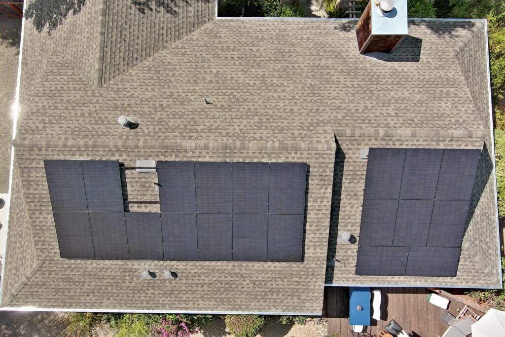Aerial view of a house roof with multiple solar panels installed on the shingles. The panels are arranged in two large rectangular groups, with chimneys and vents visible. Some greenery and part of a wooden deck are seen around the house.