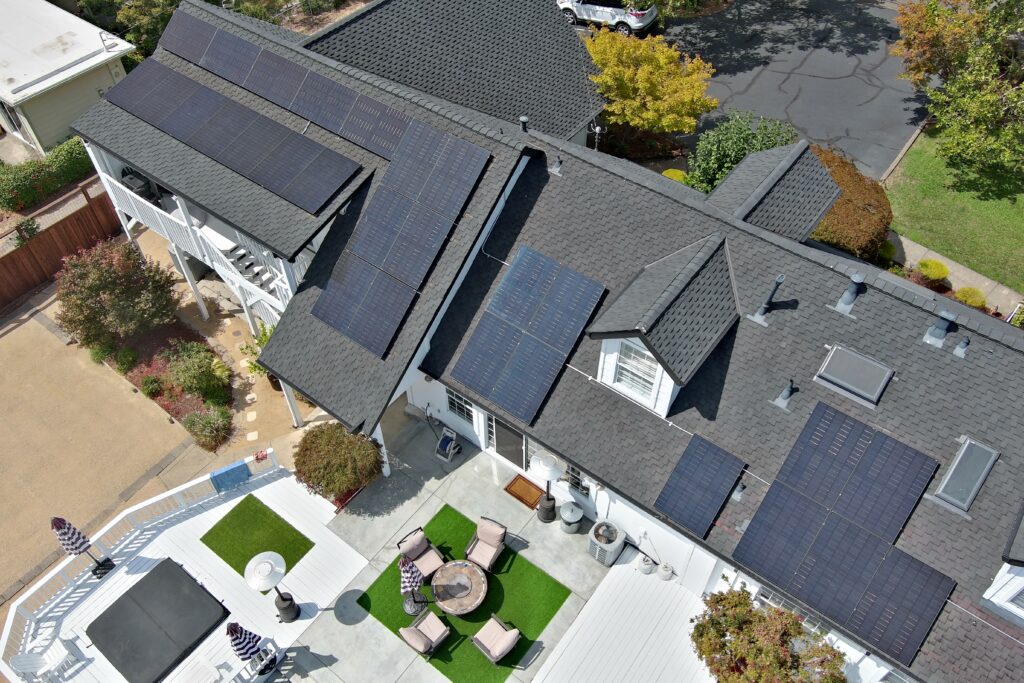 Aerial view of a house with multiple solar panels installed on its dark gray roof, surrounded by a patio, outdoor furniture, and trees.