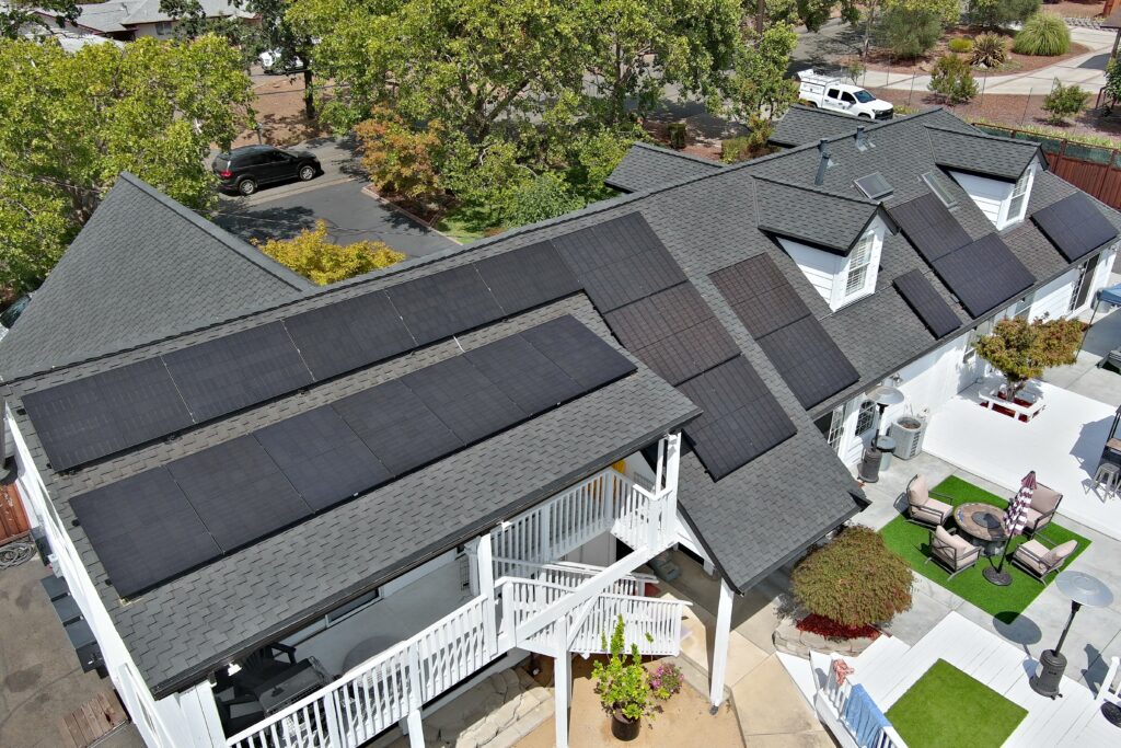 Aerial view of a house with black solar panels installed on a grey shingled roof, surrounded by trees, patio furniture, and landscaped yard.