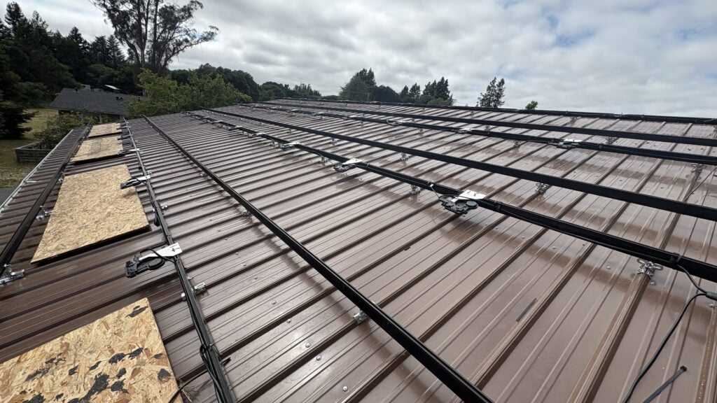 A brown metal roof with mounting rails and electrical wiring installed for solar panels, with some plywood sheets covering roof sections. Trees and a cloudy sky are in the background.