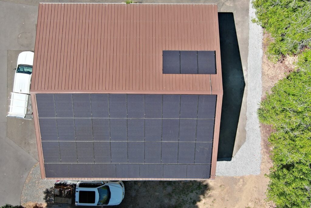 Aerial view of a building with a brown roof covered mostly by solar panels, two white vehicles parked nearby, and greenery on the right side.