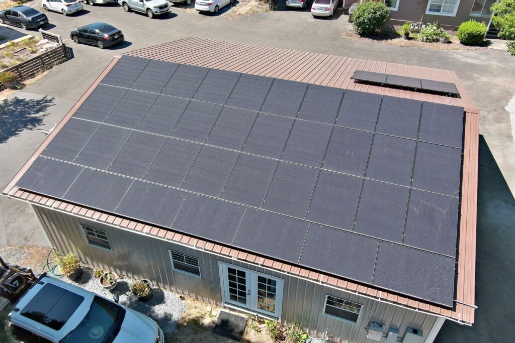 Aerial view of a metal-roofed building covered with solar panels, surrounded by parked cars and garden plants, on a sunny day.
